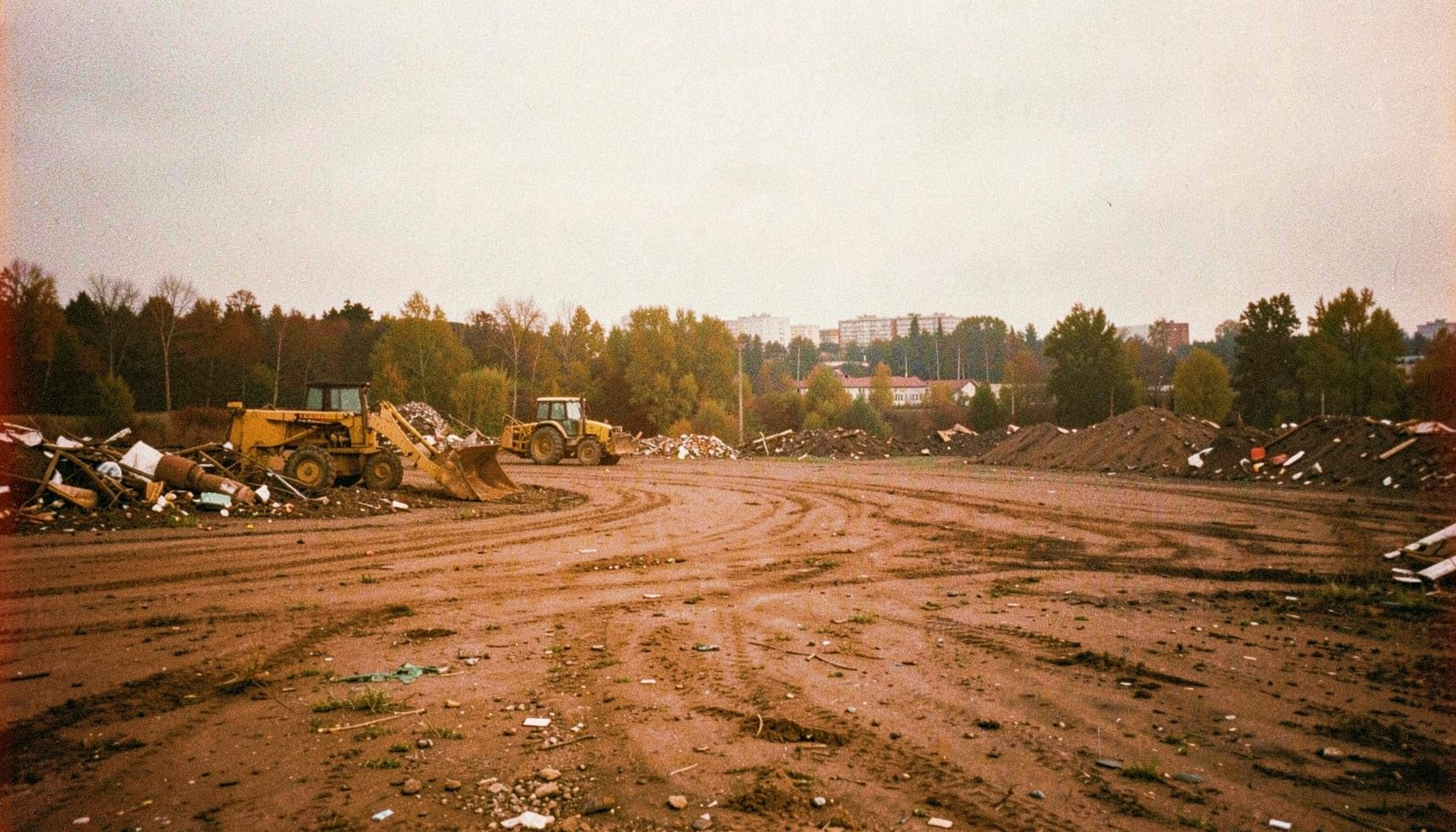 Wide panoramic view of a dirt lot or landfill site with a bulldozer and tractor, scattered debris, trees and distant buildings under an overcast sky, captured on grainy analog film